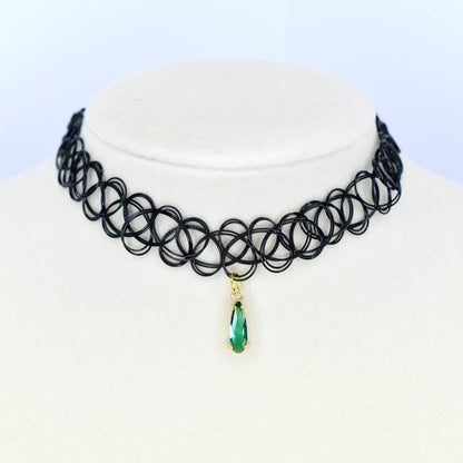 A black choker necklace with a pendant. The necklace is made of a series of interconnected black loops. A gold-colored pendant hangs from the center, featuring a teardrop-shaped green gemstone.