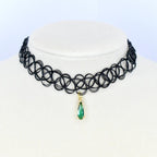 A black choker necklace with a pendant. The necklace is made of a series of interconnected black loops. A gold-colored pendant hangs from the center, featuring a teardrop-shaped green gemstone.