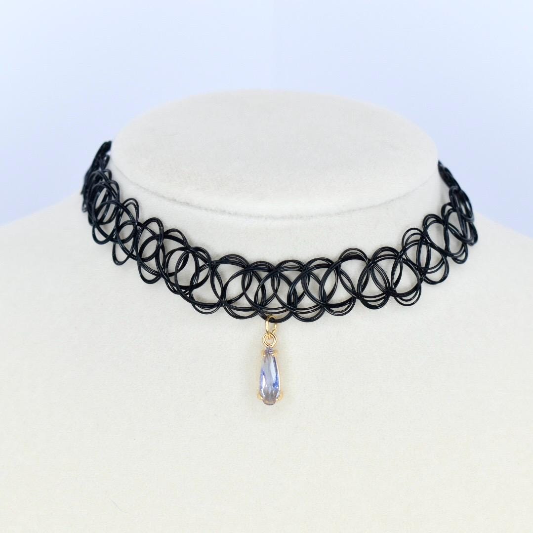 A black choker necklace with a pendant. The necklace is made of a black, stretchy material with a repeating circular design. A teardrop-shaped pendant hangs from the center, featuring a gold-colored setting and a light purple gemstone.