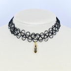 Black choker necklace with a pendant. The necklace is made of a series of interconnected black loops. A small gold-colored pendant hangs from the center featuring a teardrop-shaped dark gray gemstone. The necklace is displayed on a white mannequin.