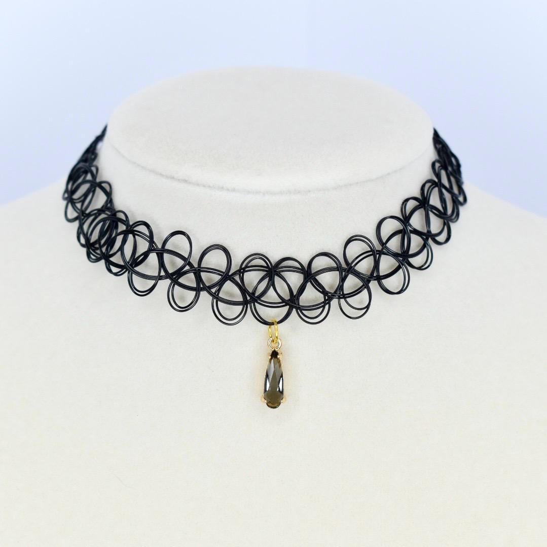 Black choker necklace with a pendant. The necklace is made of a series of interconnected black loops. A small gold-colored pendant hangs from the center featuring a teardrop-shaped dark gray gemstone. The necklace is displayed on a white mannequin.