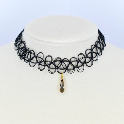 Black choker necklace with a pendant. The necklace is made of a series of interconnected black loops. A small gold-colored pendant hangs from the center featuring a teardrop-shaped dark gray gemstone. The necklace is displayed on a white mannequin.