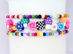Three colorful beaded bracelets. The top and bottom bracelets feature small, round beads in a rainbow of colors. The middle bracelet has flower, mushroom, and checkerboard charms. The bracelets are stacked on top of each other.
