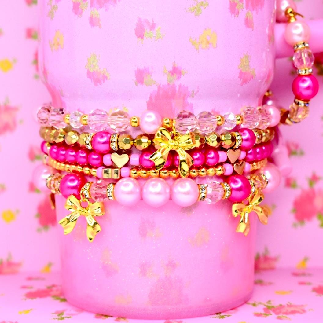 Close-up of pink tumbler bracelet stack with gold bow charms heart beads pearls and sparkly accents feminine drinkware jewelry.