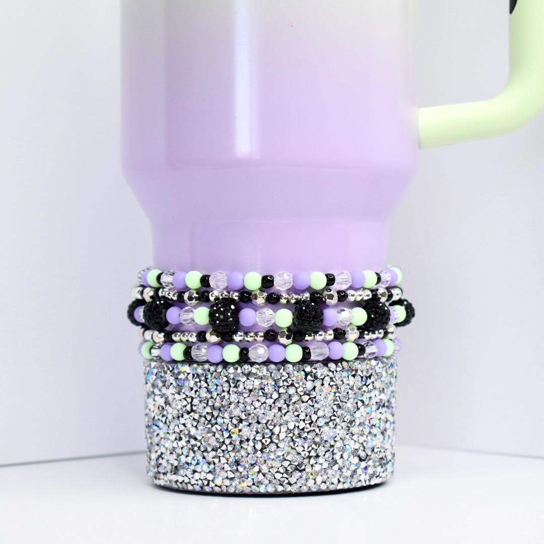 Beaded tumbler bracelet in pastel purple, mint green, black, and silver shown on a lavender tumbler with rhinestone base. Features clear faceted beads, shiny silver spacers, matte pastel beads, and textured black statement accents.