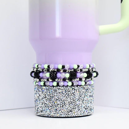 Beaded tumbler bracelet in pastel purple, mint green, black, and silver shown on a lavender tumbler with rhinestone base. Features clear faceted beads, shiny silver spacers, matte pastel beads, and textured black statement accents.