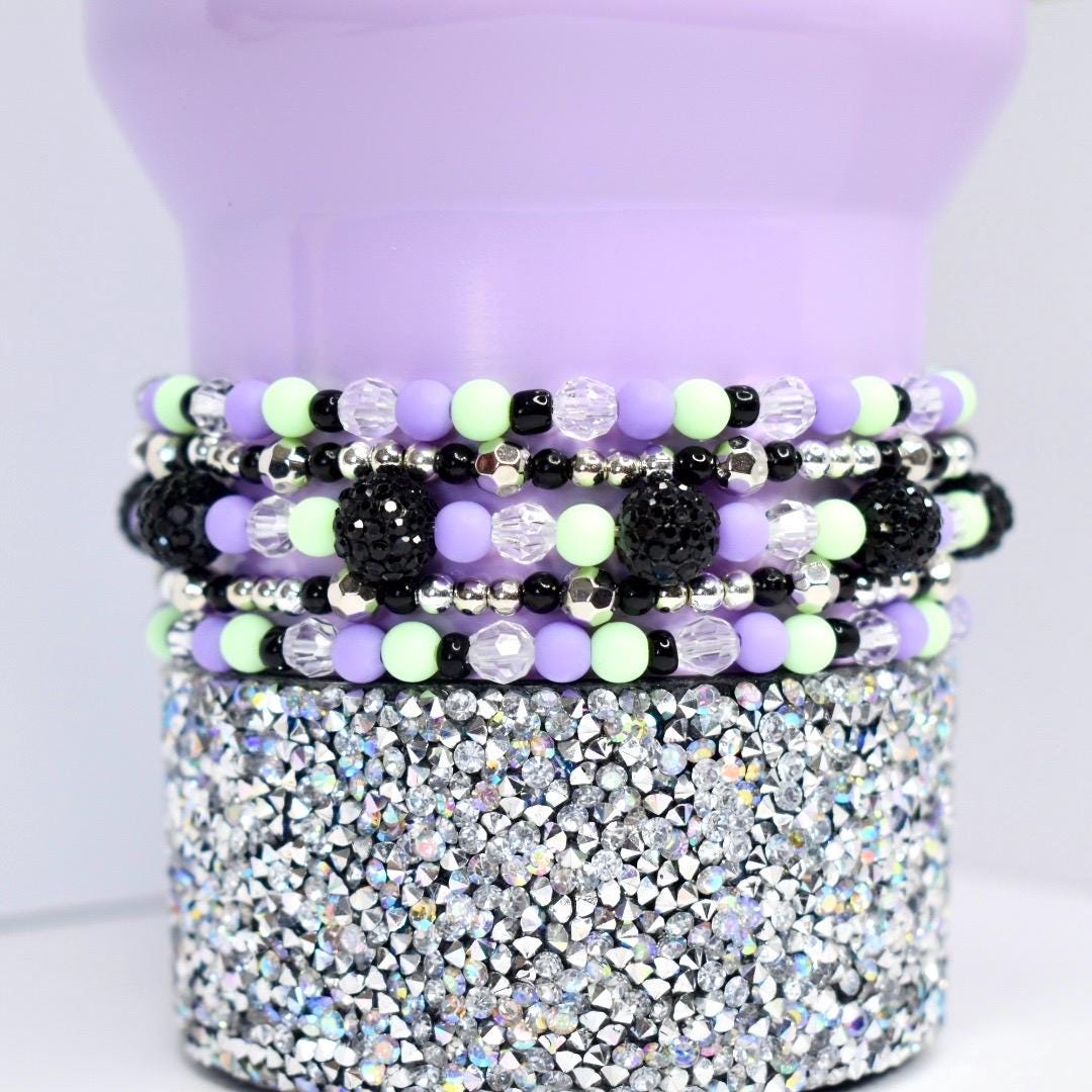 Close-up of a beaded tumbler bracelet in pastel purple, mint green, black, and silver on a lavender tumbler with a rhinestone base. Bracelet features round matte beads, faceted clear accents, shiny silver spacers, and textured black statement beads.