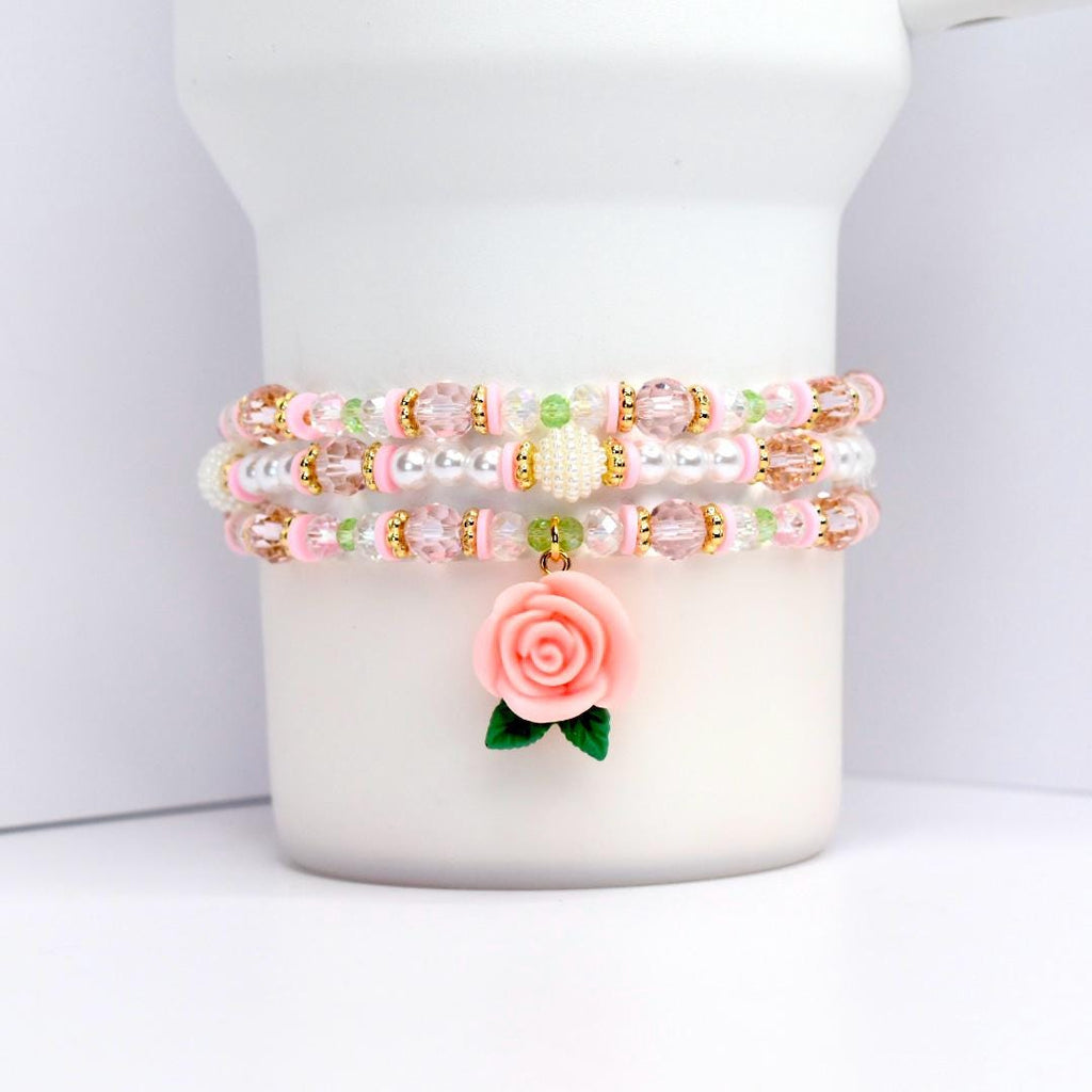 Three beaded bracelets in pink, white, and gold. Each bracelet features a combination of beads, including clear, pink, and gold. A pink rose charm with green leaves dangles from the bottom bracelet. The bracelets are displayed on a white cylindrical object.