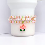 Three beaded bracelets in pink, white, and gold. Each bracelet features a combination of beads, including clear, pink, and gold. A pink rose charm with green leaves dangles from the bottom bracelet. The bracelets are displayed on a white cylindrical object.