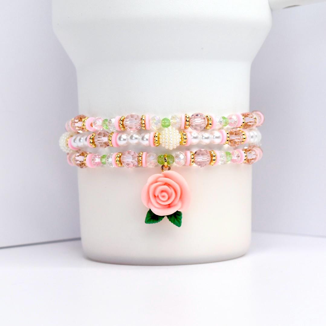 Three beaded bracelets in pink, white, and gold. Each bracelet features a combination of beads, including clear, pink, and gold. A pink rose charm with green leaves dangles from the bottom bracelet. The bracelets are displayed on a white cylindrical object.