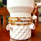 Set of five brown, gold, and white beaded tumbler bracelets wrapped around a white textured tumbler, featuring the phrase “BUT FIRST COFFEE” in letter beads and resin coffee cup charms on both the tumbler and handle bracelet.