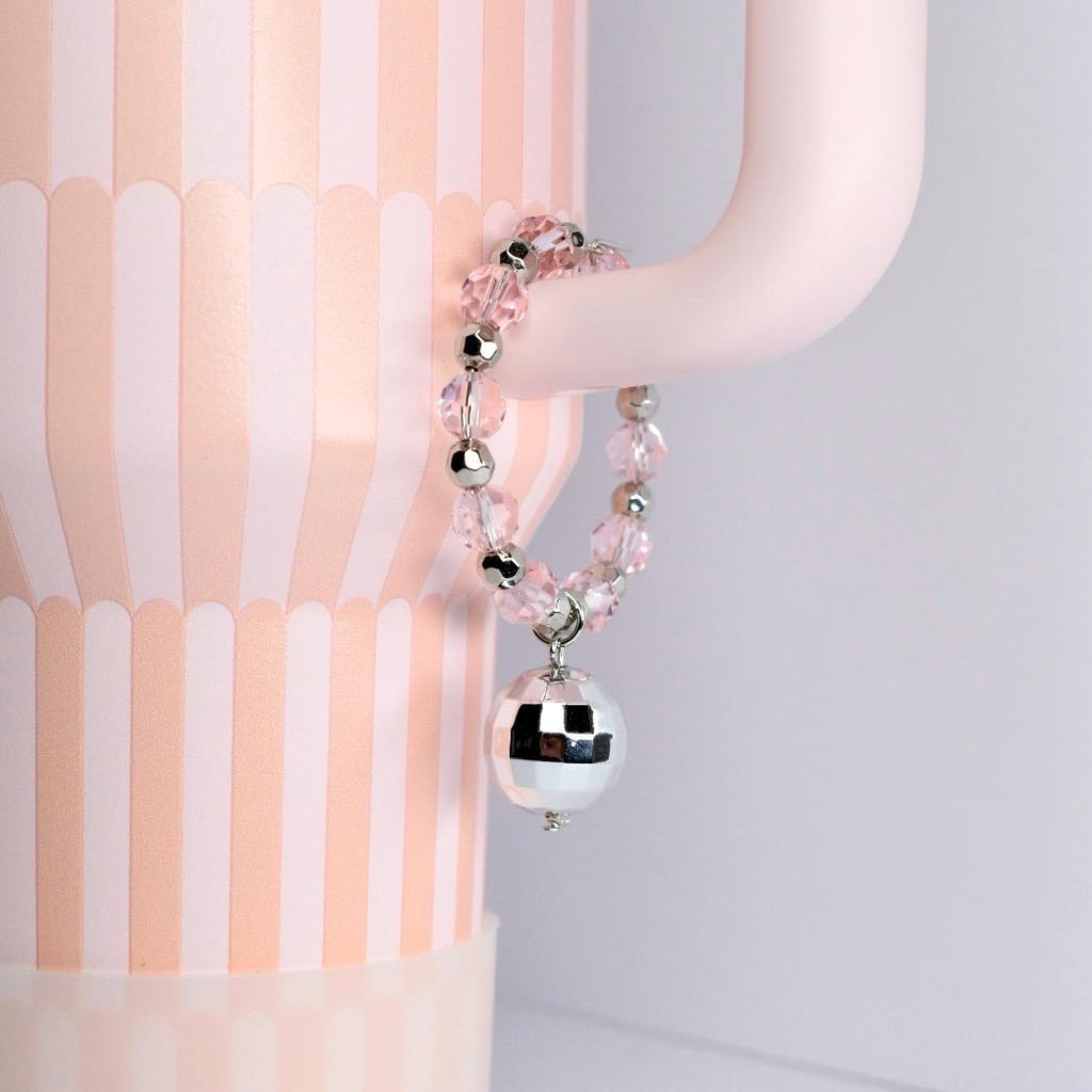 A pink beaded bracelet with silver accents and a mirrored disco ball charm. The bracelet is hanging on a light pink curved object, with a pink and white striped background.