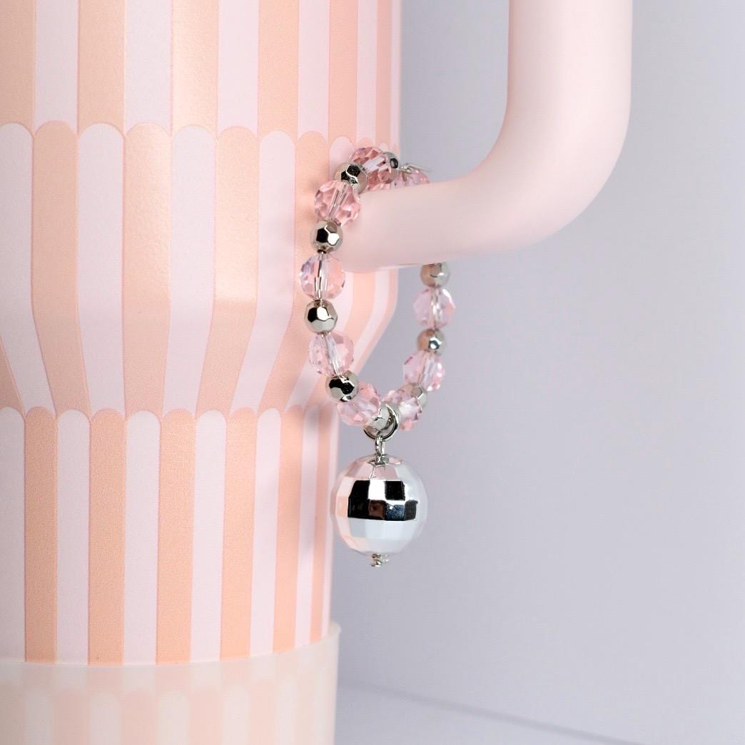 A pink beaded bracelet with silver accents and a mirrored disco ball charm. The bracelet is hanging on a light pink curved object, with a pink and white striped background.