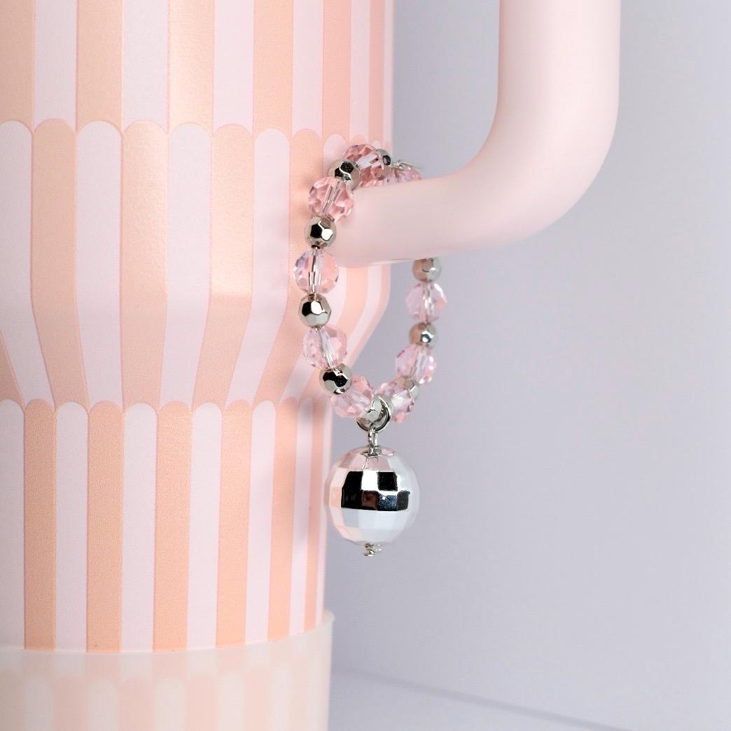 A pink beaded bracelet with silver accents and a mirrored disco ball charm. The bracelet is hanging on a light pink curved object, with a pink and white striped background.