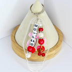 A clear acrylic keychain with the word 'OFFICE' spelled out in white letter beads. The keychain features red and white beads, a pink ribbon, and a cherry charm. The keychain is attached to a silver keyring.