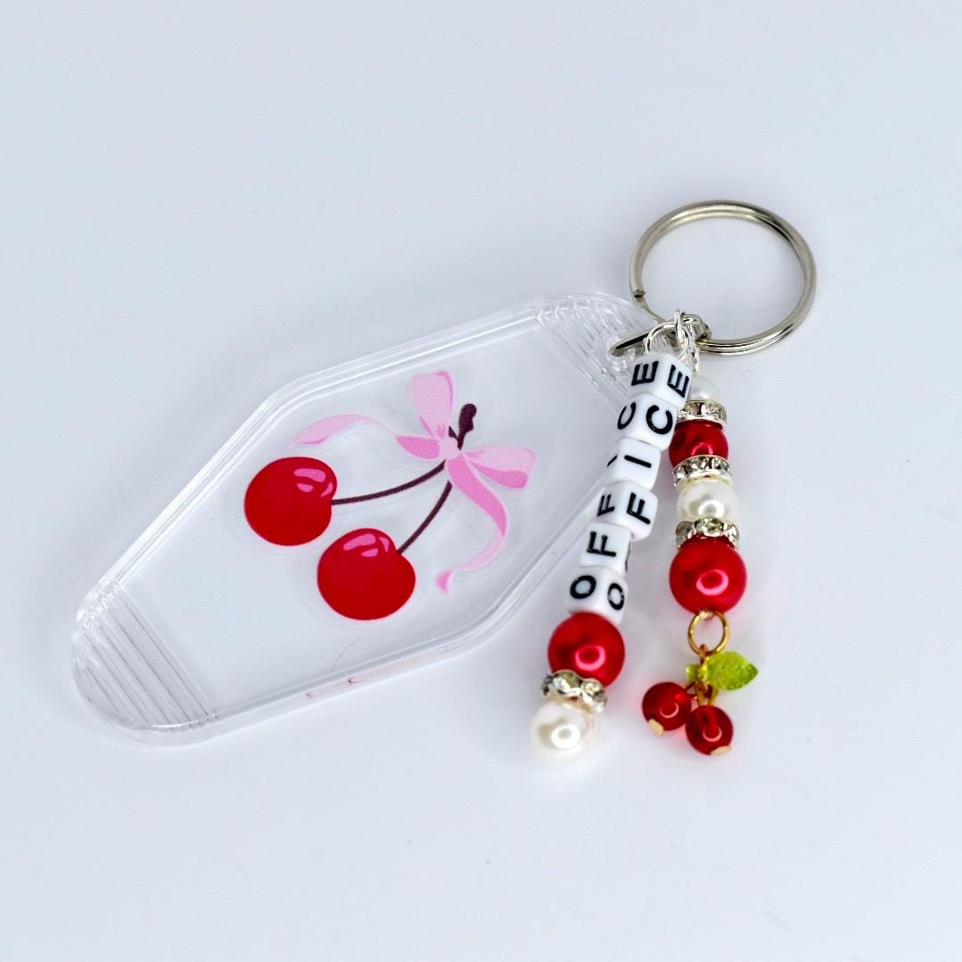 A clear, plastic keychain with a cherry design. The keychain features a pink bow and two red cherries. The keychain also has a metal ring and a beaded charm that spells 'OFFICE' in white letters. The charm includes red beads and faux pearls.