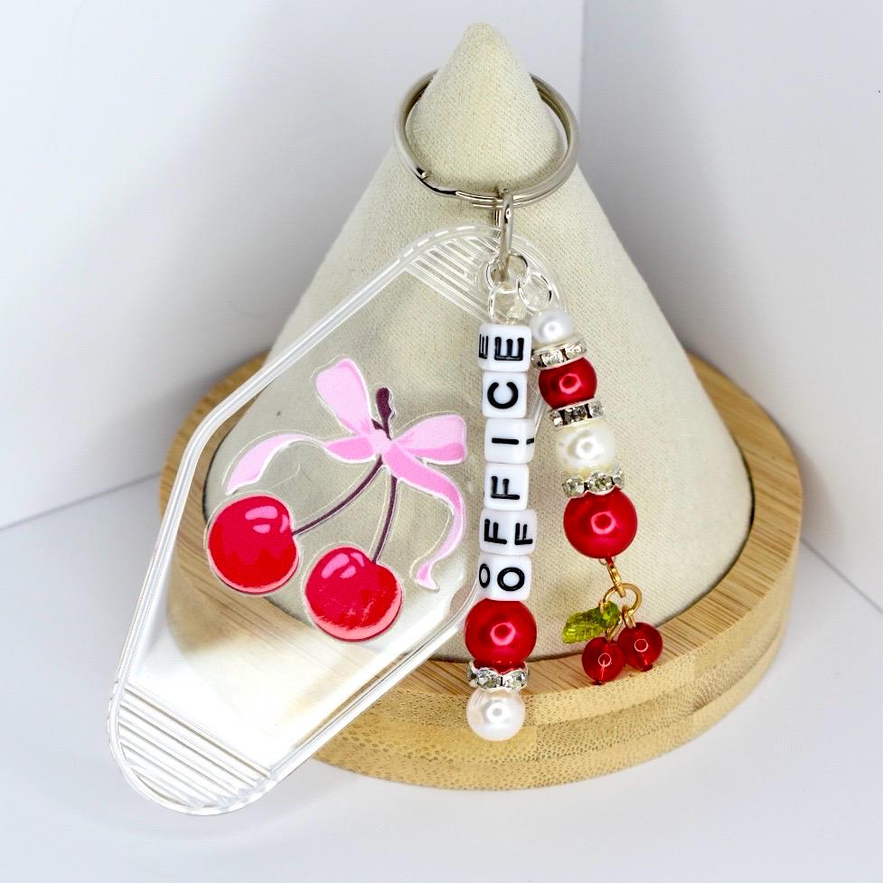 A clear acrylic keychain with a cherry design. The keychain features a painted image of red cherries with a pink bow. The word 'OFFICE' is spelled out with white letter beads. Additional red beads, white pearls, and a small cherry charm are attached to the keychain.