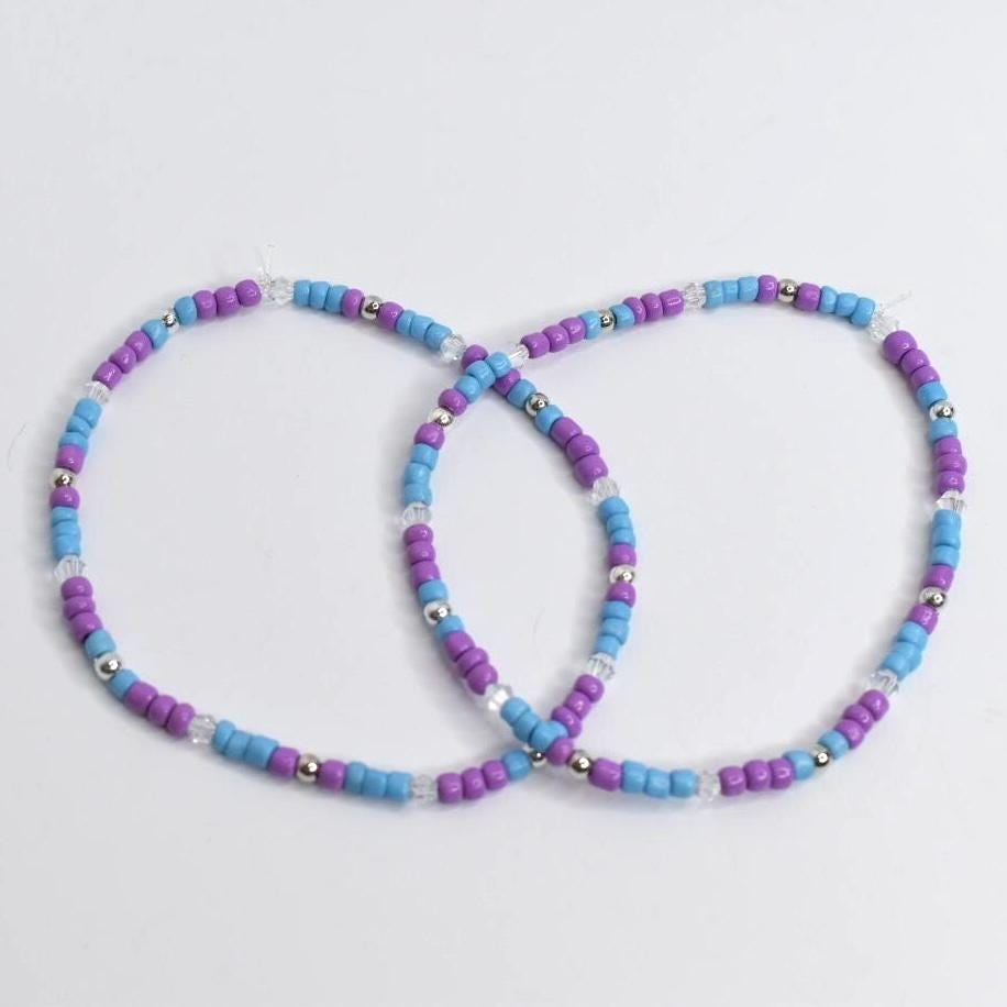 Two beaded bracelets. Each bracelet features a repeating pattern of purple, light blue, and clear beads, interspersed with silver-colored beads. The bracelets are circular and appear to be handmade, suitable for casual wear.
