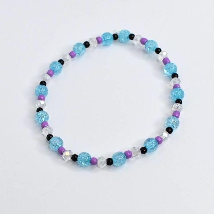 A beaded bracelet featuring light blue, purple, black, and clear beads. The bracelet has a mix of round and faceted beads, creating a colorful and textured accessory. The bracelet is on a white background.