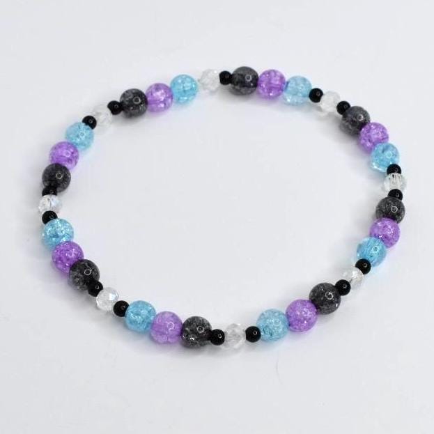 A beaded bracelet featuring a variety of colorful beads. The bracelet includes light blue, purple, black, and clear beads. The beads are strung together to form a circular shape. The bracelet is on a white background.