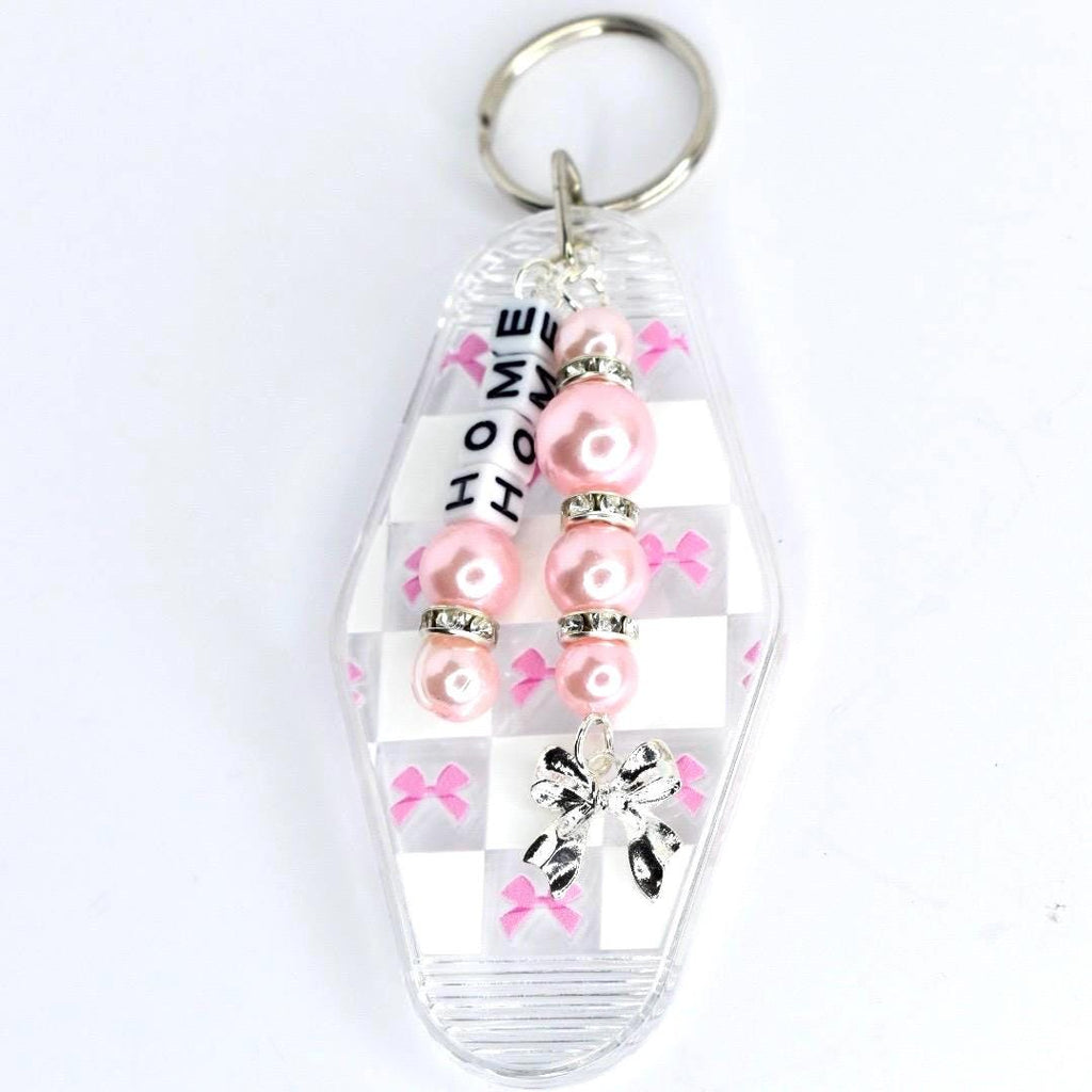 A clear, rectangular keychain with a pink and white checkered pattern. The keychain has a silver key ring at the top and a charm with pink beads and the word 'HOME' spelled out in white letters. A silver bow charm hangs at the bottom.