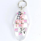 A clear, rectangular keychain with a pink and white checkered pattern. The keychain has a silver key ring at the top and a charm with pink beads and the word 'HOME' spelled out in white letters. A silver bow charm hangs at the bottom.