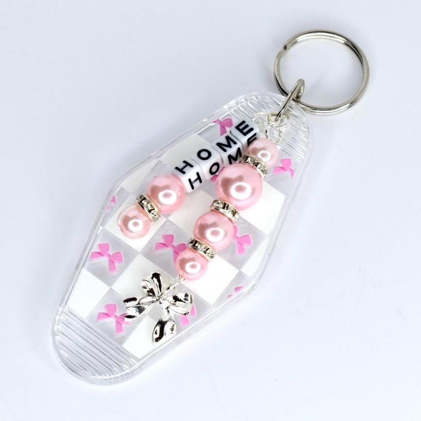 A clear motel-style keychain with a silver ring. The keychain has a white and pink checkered pattern. It features pink pearl beads with silver rhinestone accents and letter beads spelling 'HOME HOME'. Pink bow accents and a silver bow charm complete the design.