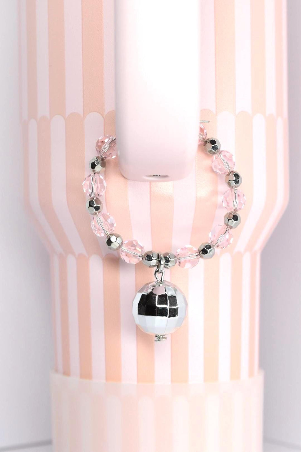 A pink and silver beaded bracelet with a small disco ball charm. The bracelet is made of clear and silver beads. The disco ball charm is silver and reflective. The background is a pink and white striped pattern.