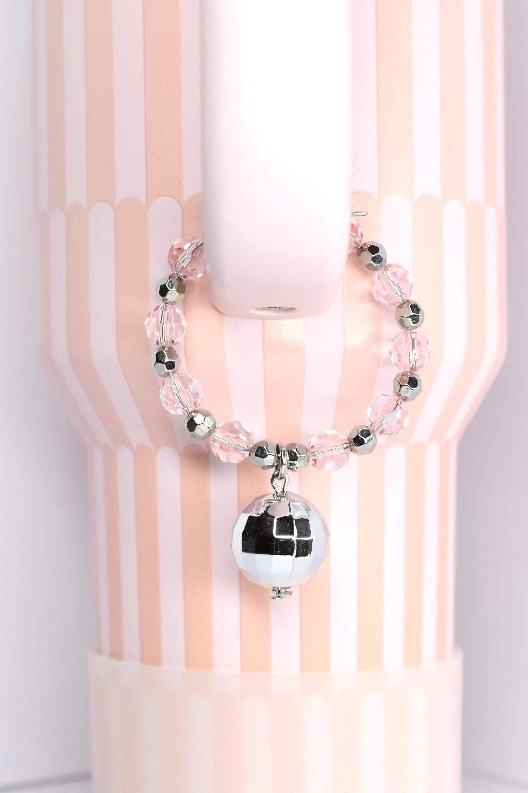 A pink and silver beaded bracelet with a small disco ball charm. The bracelet is made of clear and silver beads. The disco ball charm is silver and reflective. The background is a pink and white striped pattern.