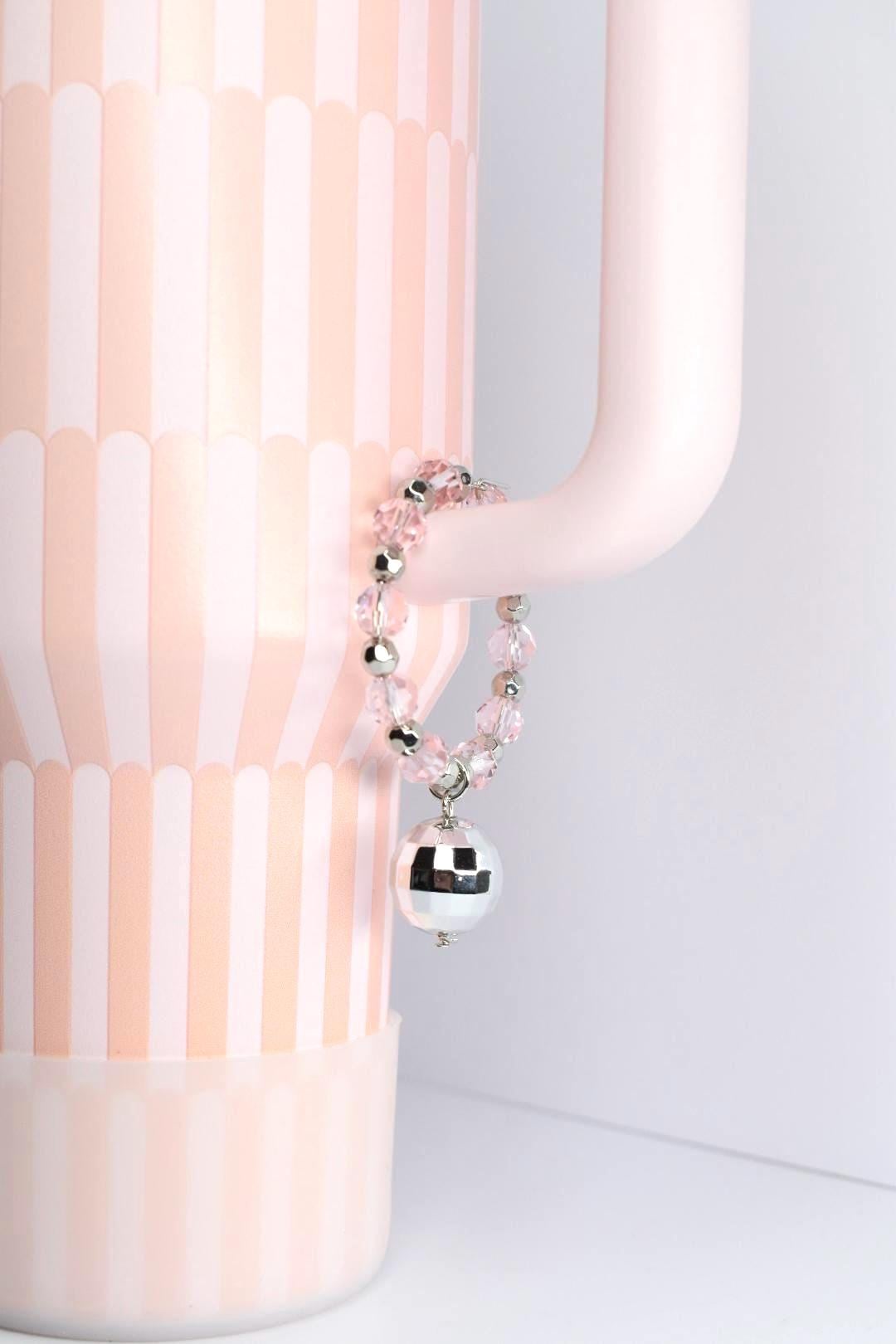 A pink and white Stanley tumbler with a handle bracelet. The bracelet features pink beads, silver accents, and a mirrored disco ball charm. The overall aesthetic is playful and decorative.