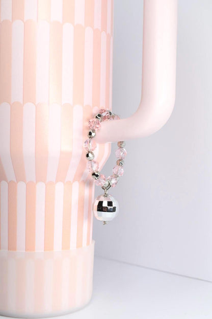 A pink and white Stanley tumbler with a handle bracelet. The bracelet features pink beads, silver accents, and a mirrored disco ball charm. The overall aesthetic is playful and decorative.