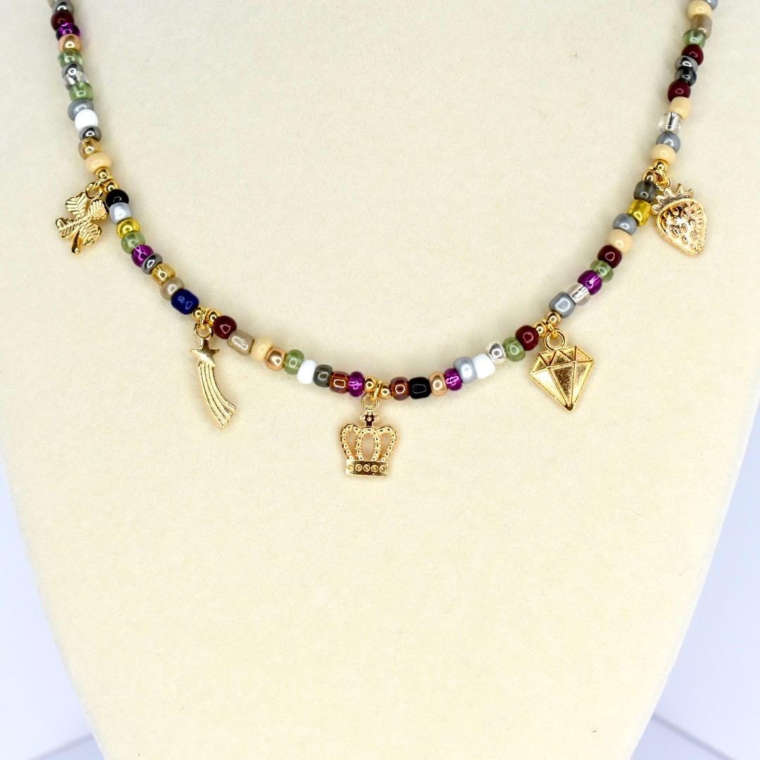 A beaded necklace with a variety of colorful beads and gold-colored charms. The charms include a crown, a diamond, a shooting star, a strawberry, and a four-leaf clover. The necklace is displayed on a white surface.