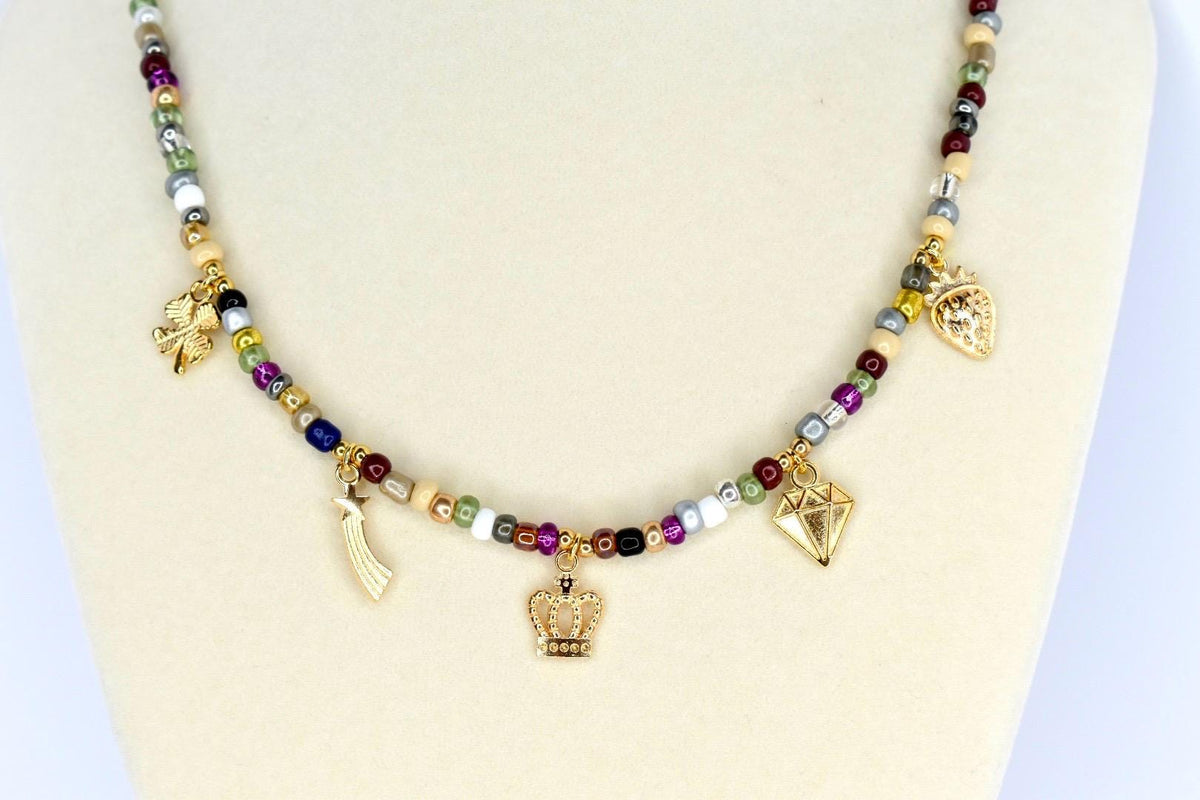 A beaded necklace with a variety of colorful beads and gold-colored charms. The charms include a four-leaf clover, a shooting star, a crown, a diamond, and a strawberry. The necklace is displayed on a white mannequin.