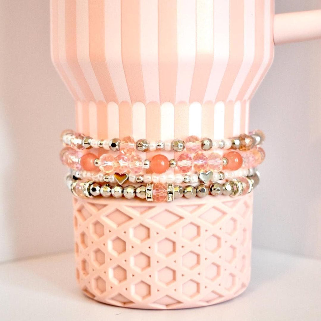 A set of four beaded tumbler bracelets wrapped around a blush pink Stanley-style tumbler with a textured base. The bracelets feature coral, blush, crystal, and silver beads with accents like metallic hearts and decorative spacers, creating a soft, romantic look.