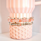 A set of four beaded tumbler bracelets wrapped around a blush pink Stanley-style tumbler with a textured base. The bracelets feature coral, blush, crystal, and silver beads with accents like metallic hearts and decorative spacers, creating a soft, romantic look.