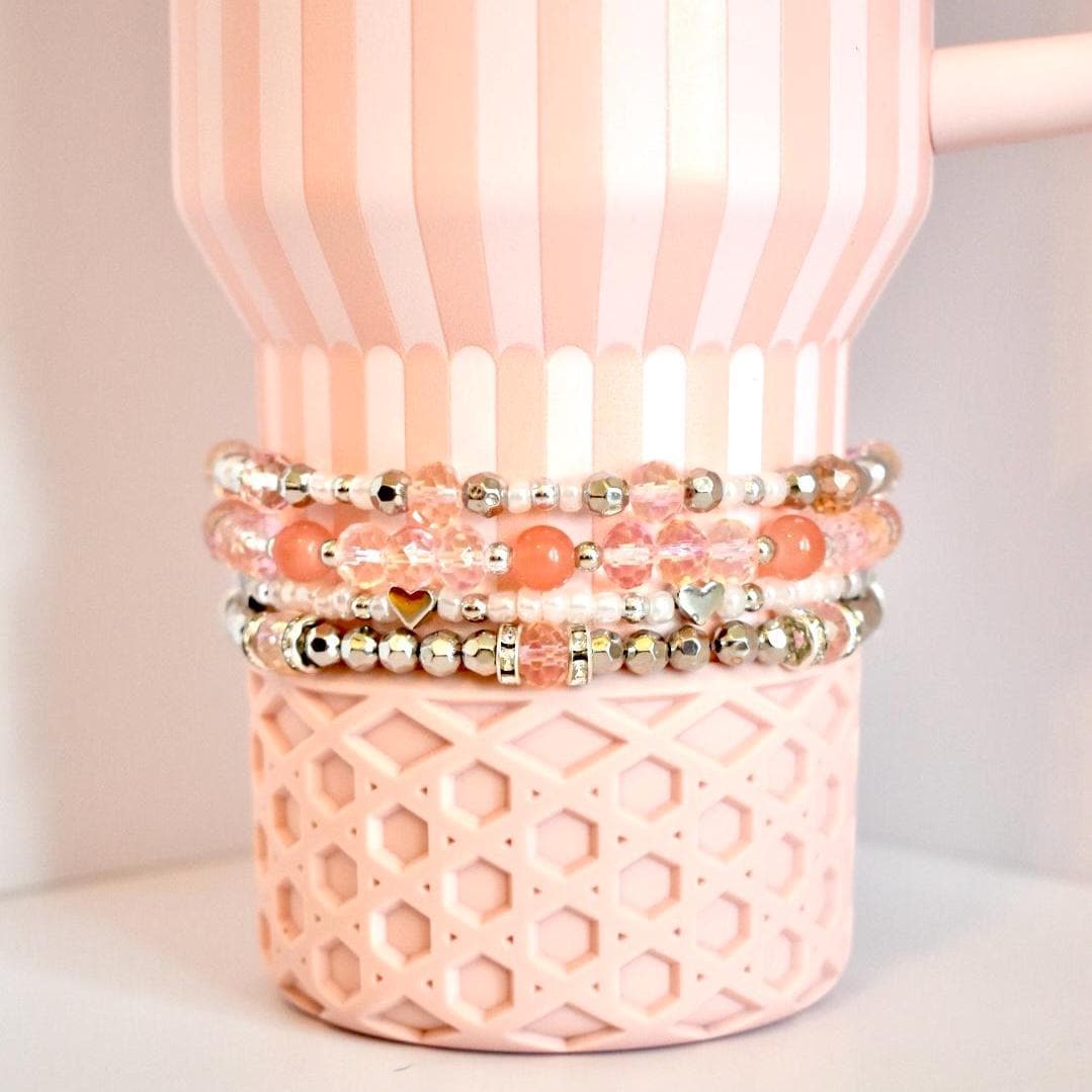 A set of four beaded tumbler bracelets wrapped around a blush pink Stanley-style tumbler with a textured base. The bracelets feature coral, blush, crystal, and silver beads with accents like metallic hearts and decorative spacers, creating a soft, romantic look.