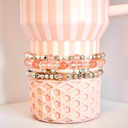 A set of four beaded tumbler bracelets wrapped around a blush pink Stanley-style tumbler with a textured base. The bracelets feature coral, blush, crystal, and silver beads with accents like metallic hearts and decorative spacers, creating a soft, romantic look.