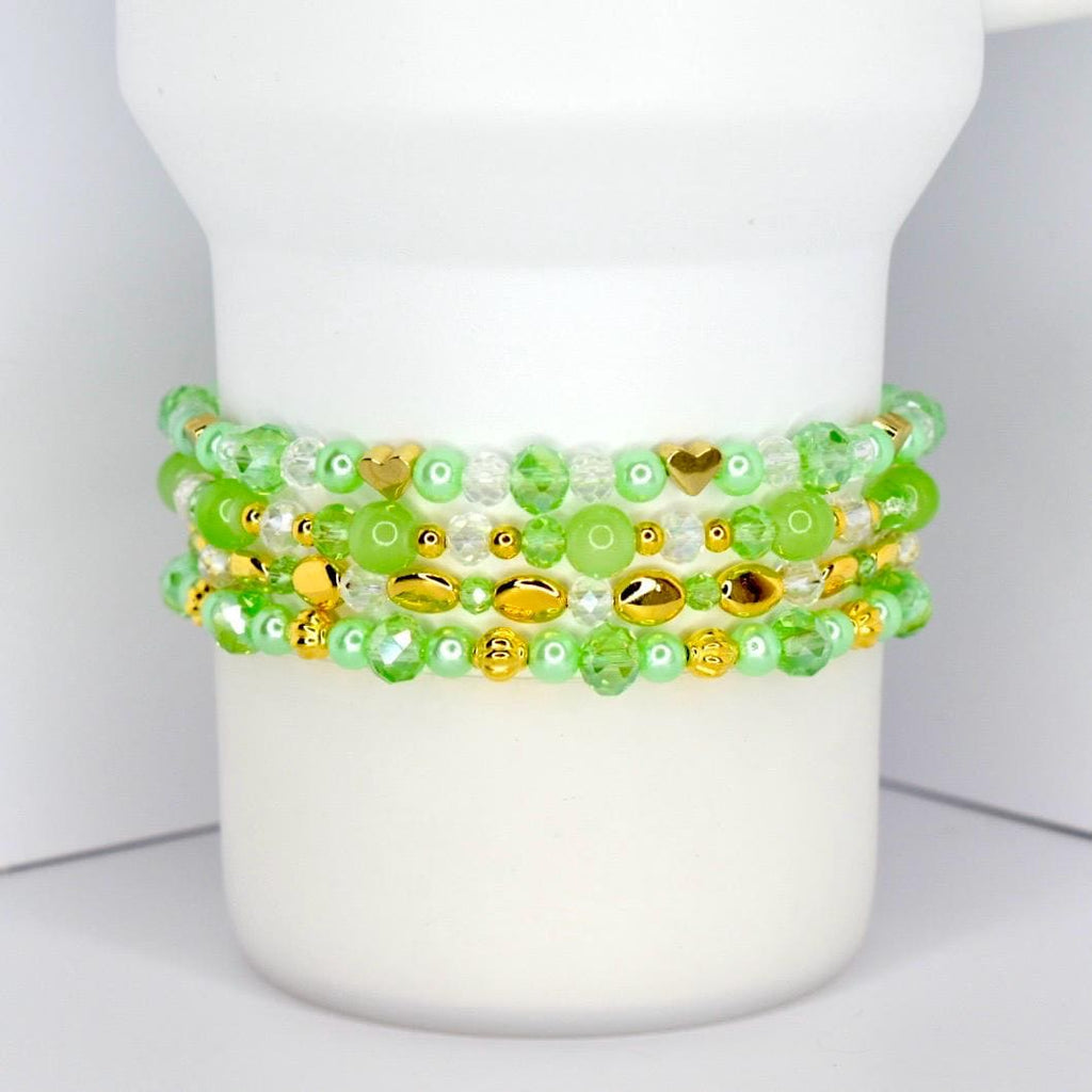Four beaded bracelets in shades of green and gold are stacked on a white cylindrical object. The bracelets feature various bead shapes, including round, oval, and heart-shaped beads. The beads are a mix of clear, opaque, and metallic gold.