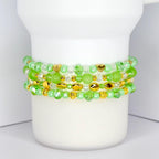 Four beaded bracelets in shades of green and gold are stacked on a white cylindrical object. The bracelets feature various bead shapes, including round, oval, and heart-shaped beads. The beads are a mix of clear, opaque, and metallic gold.