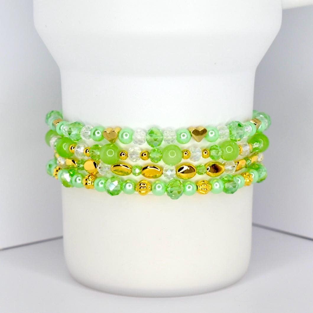 Four beaded bracelets in shades of green and gold are stacked on a white cylindrical object. The bracelets feature various bead shapes, including round, oval, and heart-shaped beads. The beads are a mix of clear, opaque, and metallic gold.