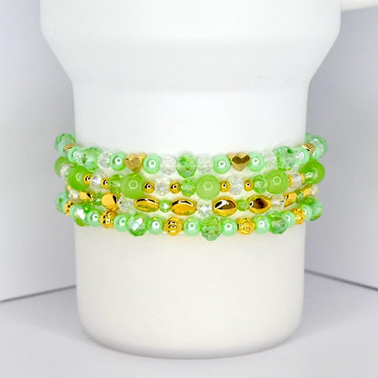 Four beaded bracelets in shades of green and gold are stacked on a white cylindrical object. The bracelets feature various bead shapes, including round, oval, and heart-shaped beads. The beads are a mix of clear, opaque, and metallic gold.