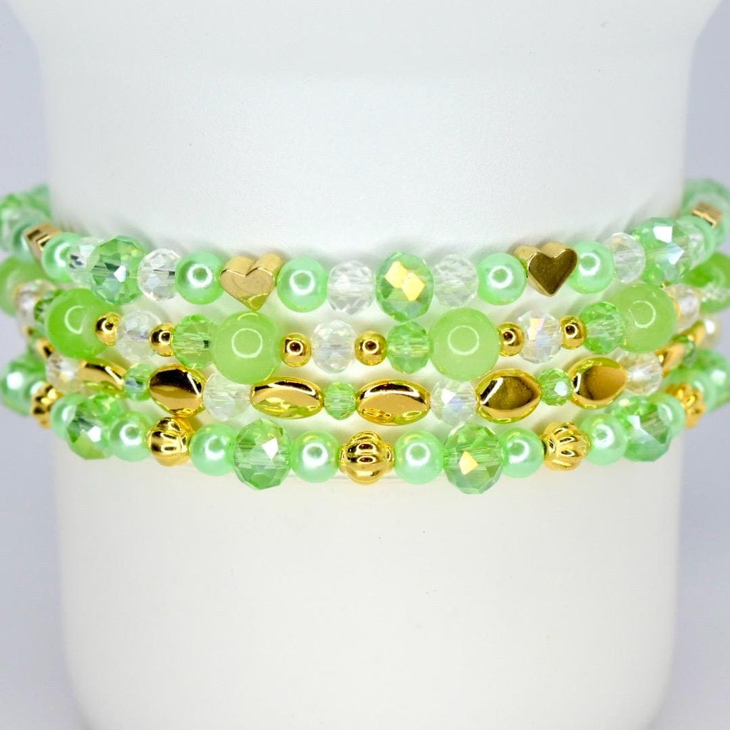 Four beaded bracelets in shades of green and gold. The bracelets feature a variety of beads, including round, faceted, and heart-shaped beads. The bracelets are stacked on top of each other.