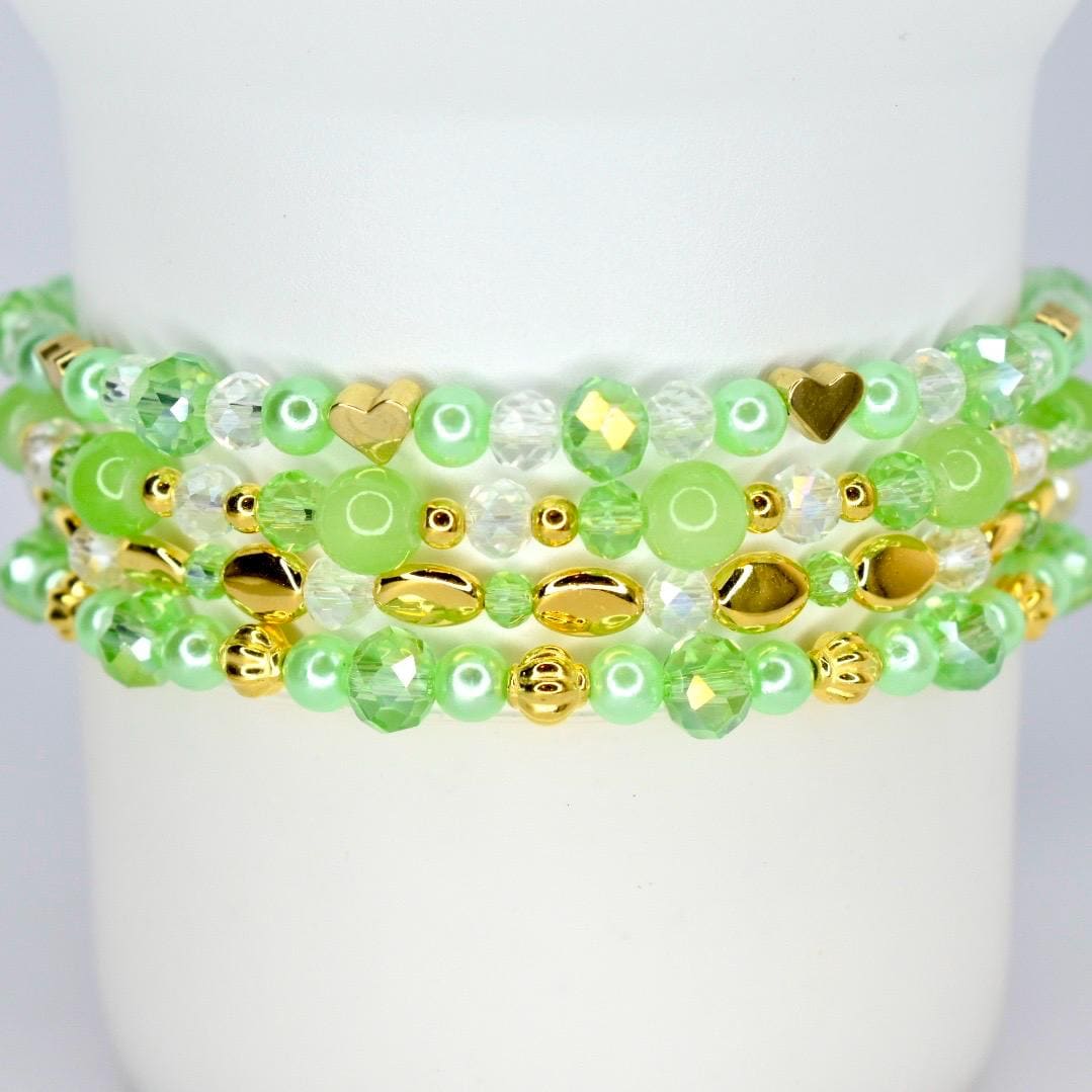 Four beaded bracelets in shades of green and gold. The bracelets feature a variety of beads, including round, faceted, and heart-shaped beads. The bracelets are stacked on top of each other.