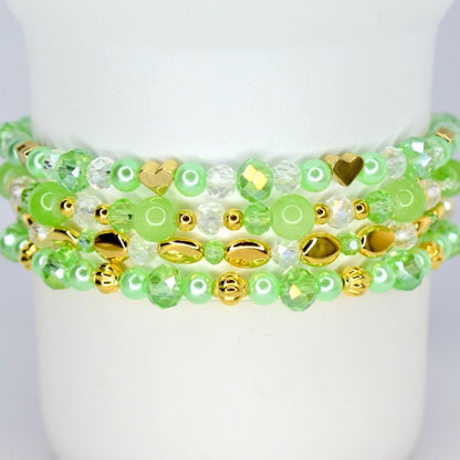 Four beaded bracelets in shades of green and gold. The bracelets feature a variety of beads, including round, faceted, and heart-shaped beads. The bracelets are stacked on top of each other.