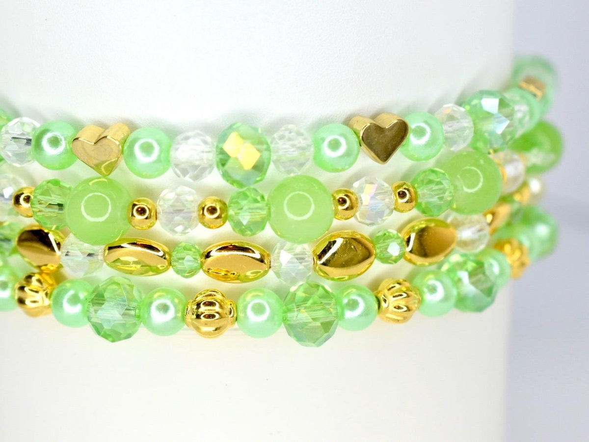 Close-up of three stacked bracelets with green, gold, and clear beads. The bracelets feature a variety of bead shapes, including round, faceted, and heart-shaped beads. The color palette includes shades of green, gold, and clear.