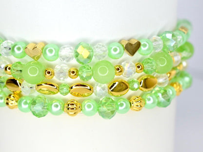 Close-up of three stacked bracelets with green, gold, and clear beads. The bracelets feature a variety of bead shapes, including round, faceted, and heart-shaped beads. The color palette includes shades of green, gold, and clear.