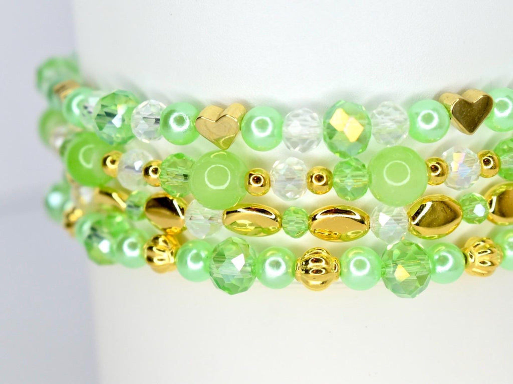 Close-up of several beaded bracelets. The bracelets feature a mix of light green, clear, and gold beads. Some beads are heart-shaped or faceted, adding texture and visual interest. The bracelets are stacked, showcasing the design.