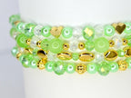 Close-up of several beaded bracelets. The bracelets feature a mix of light green, clear, and gold beads. Some beads are heart-shaped or faceted, adding texture and visual interest. The bracelets are stacked, showcasing the design.