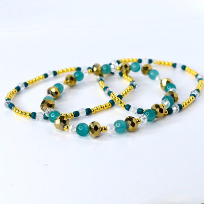 Gold, teal, and clear beaded tumbler bracelet laid flat to show full length. Features faceted metallic gold beads, smooth teal beads, and sparkling clear accents with gold spacer beads.