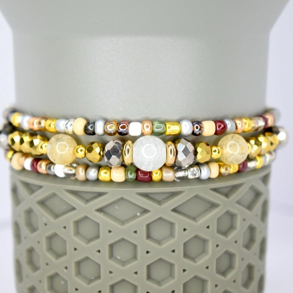 Three beaded bracelets with a variety of colors and textures. The bracelets feature gold, silver, and bronze beads, along with larger, round, and faceted beads. The bracelets are stacked on top of a gray, textured surface.